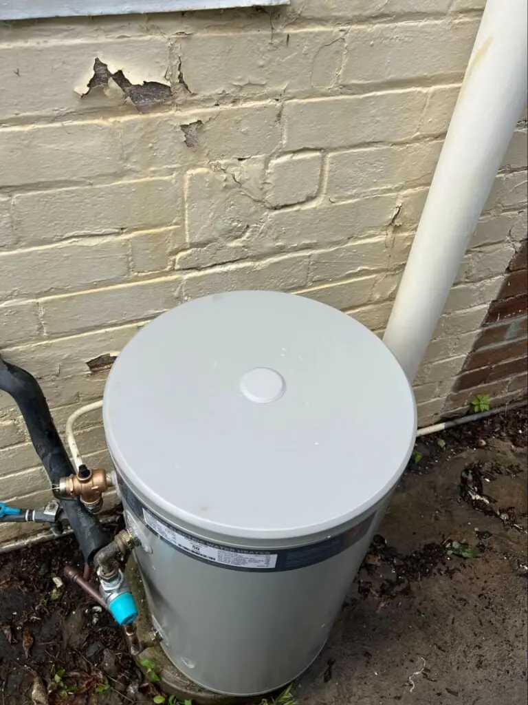 Old electric hot water unit beside a brick wall before its replacement